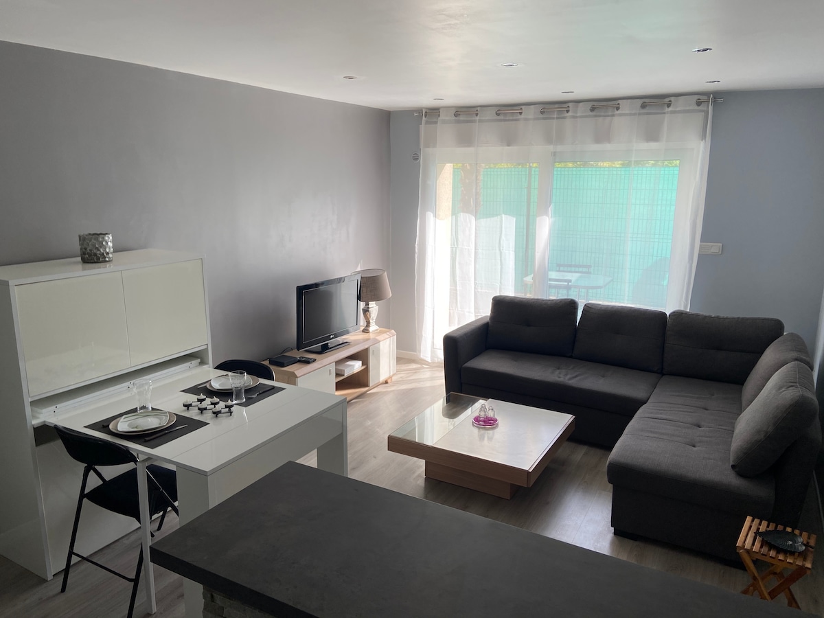 A modern living space is presented, featuring a sectional sofa and a coffee table. A flat-screen television is positioned against the wall. The dining area includes a table and chairs, while large windows allow natural light to brighten the room, with a view of a terrace.