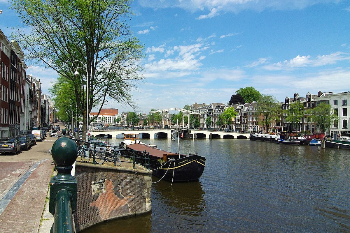 A peaceful canal scene features tree-lined pathways alongside still waters. Historic buildings rise above the water, with bridges arching gracefully across the canal. Boats are moored along the banks, and soft blue skies with scattered clouds create a serene backdrop.