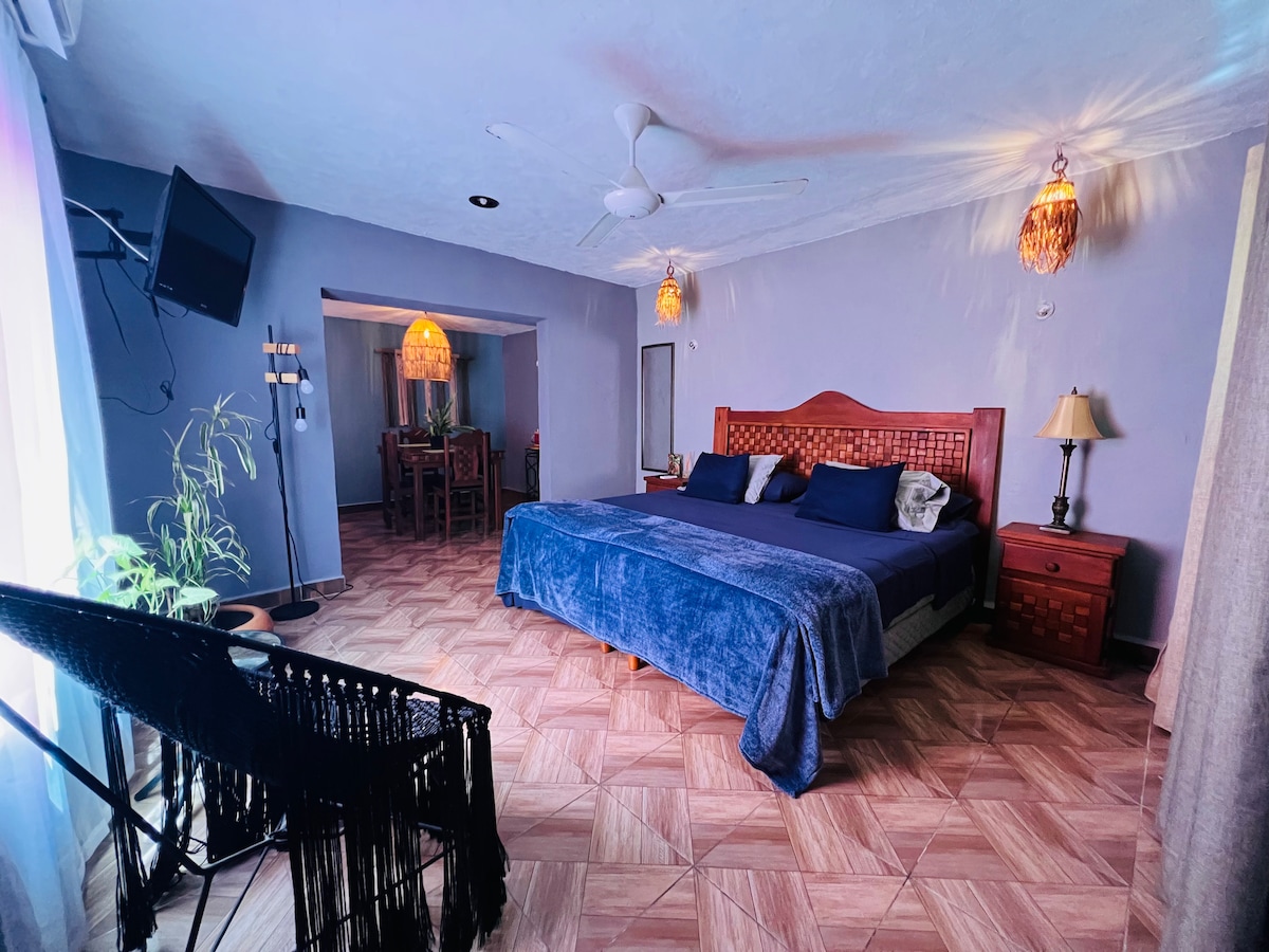 A spacious room features a king-size bed dressed in a deep blue blanket, accompanied by decorative pillows. Soft lighting is provided by two hanging fixtures. A small bedside lamp and a dark wooden nightstand are present. A seating area with a chair and a large window allows natural light to enter.
