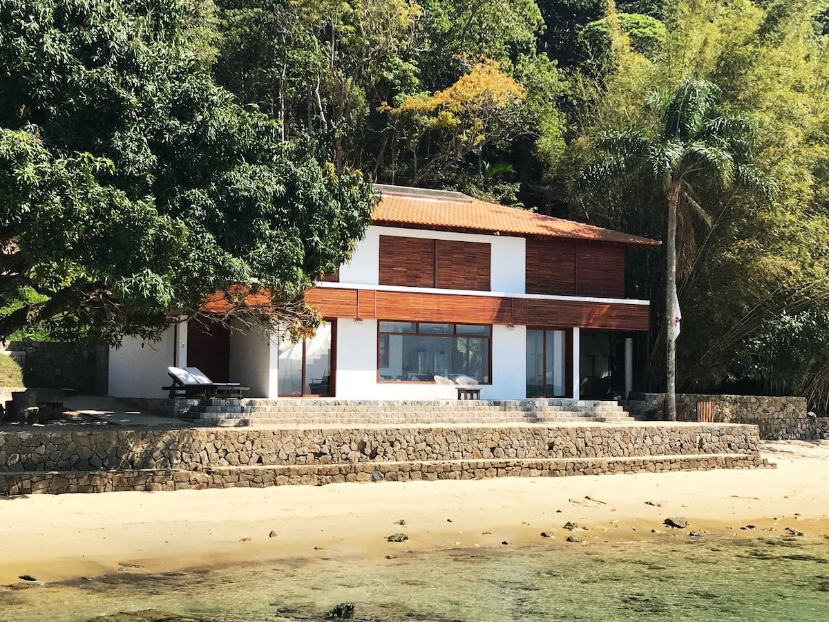 A beachfront house is presented with wooden paneling and large windows that offer views of the surrounding greenery. The structure is positioned on a stone foundation, complemented by a sandy area leading to the water's edge. Trees provide shade alongside the property.
