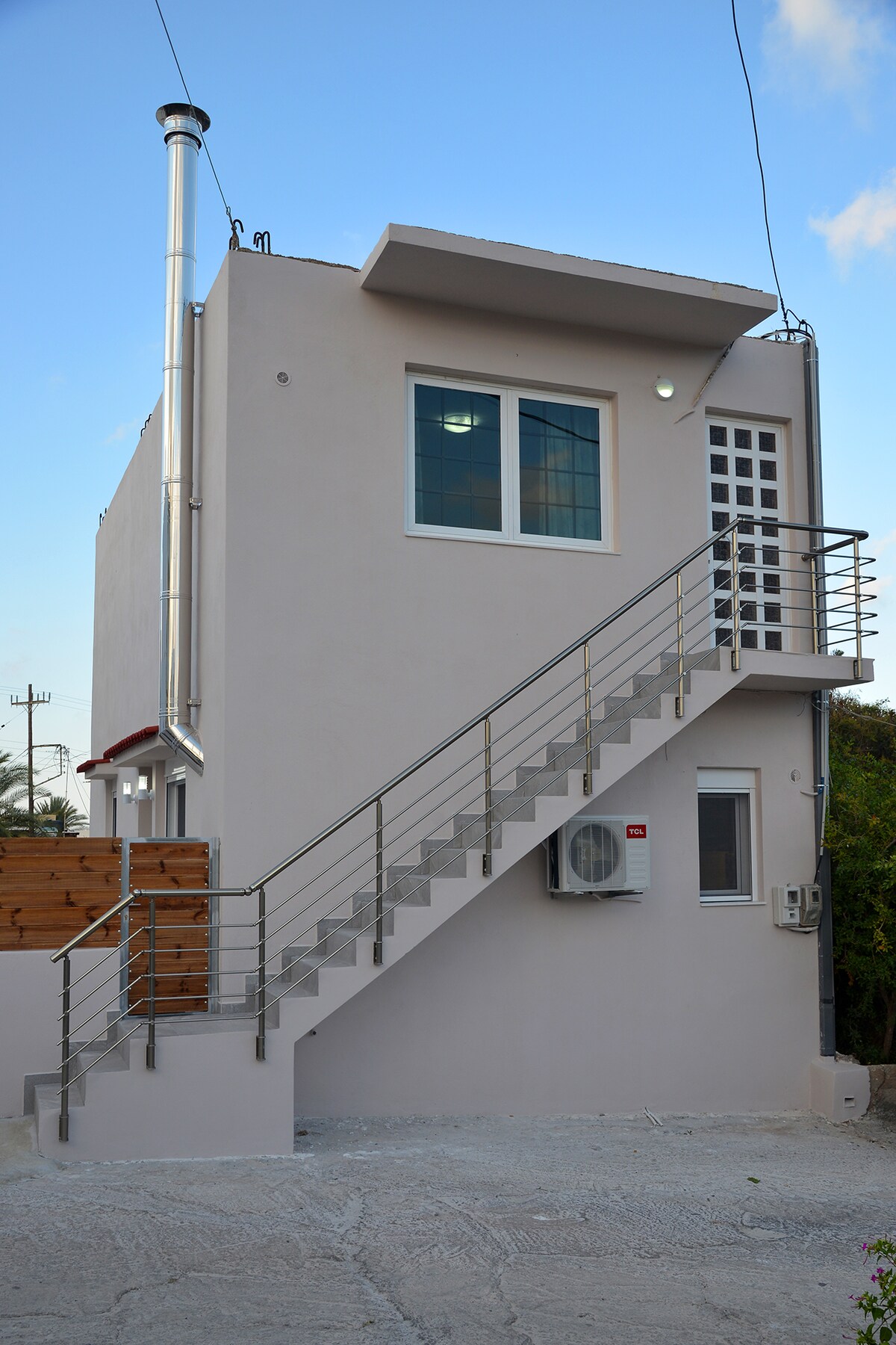 A modern, two-story house is presented, featuring a light exterior and a prominent staircase with a metal railing. Large windows are visible on both levels, providing ample light. An air conditioning unit is installed on the side, and nearby greenery adds a natural touch.