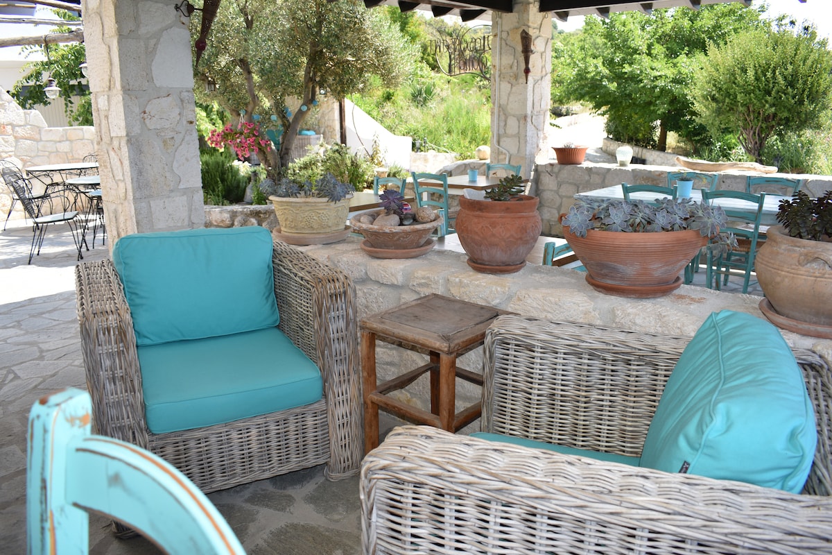 A relaxing outdoor seating area is presented with woven wicker chairs featuring soft turquoise cushions. A wooden side table is positioned between the chairs, surrounded by terracotta pots filled with various green plants. The stone structure adds a rustic charm, with greenery visible in the background.
