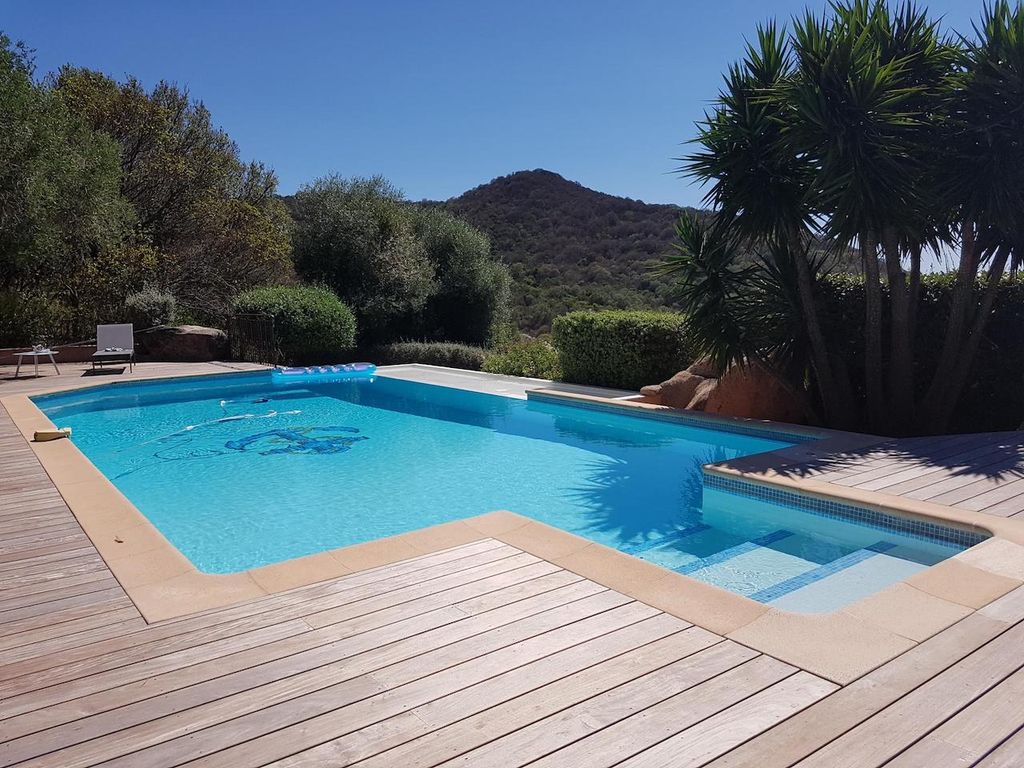 A tranquil swimming pool surrounded by a wooden deck features clear blue water, inviting relaxation. Lush greenery and shrubs frame the area, enhancing the natural setting. A distant hill is visible, adding to the serene atmosphere.