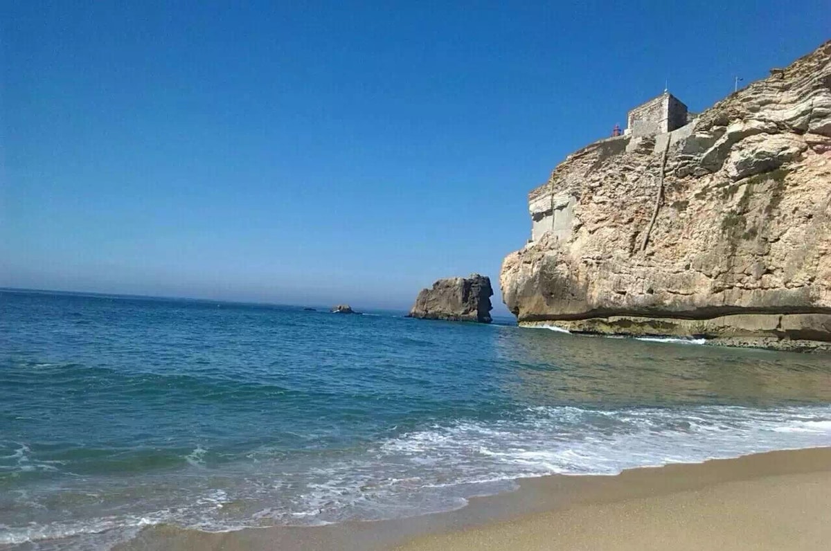 A view of a sandy beach with gentle waves lapping at the shore. A rocky cliff rises in the background, adorned with a small structure. Clear blue skies stretch overhead, complementing the serene waters.