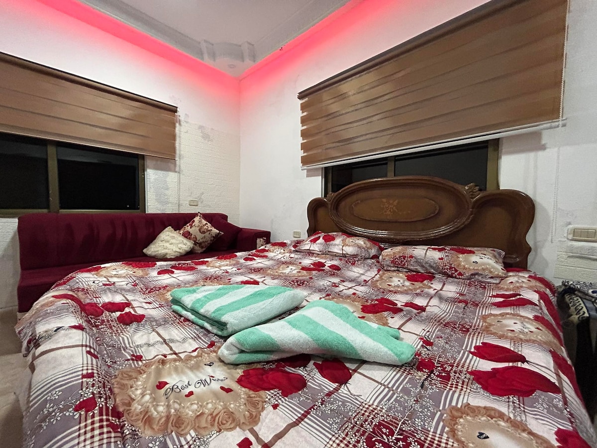 A sleeping area features a spacious bed dressed in a floral-patterned coverlet. Two towels are neatly placed at the foot of the bed. A cozy seating area with a maroon couch is positioned against the wall. Soft ambient lighting enhances the room's tranquil feel.