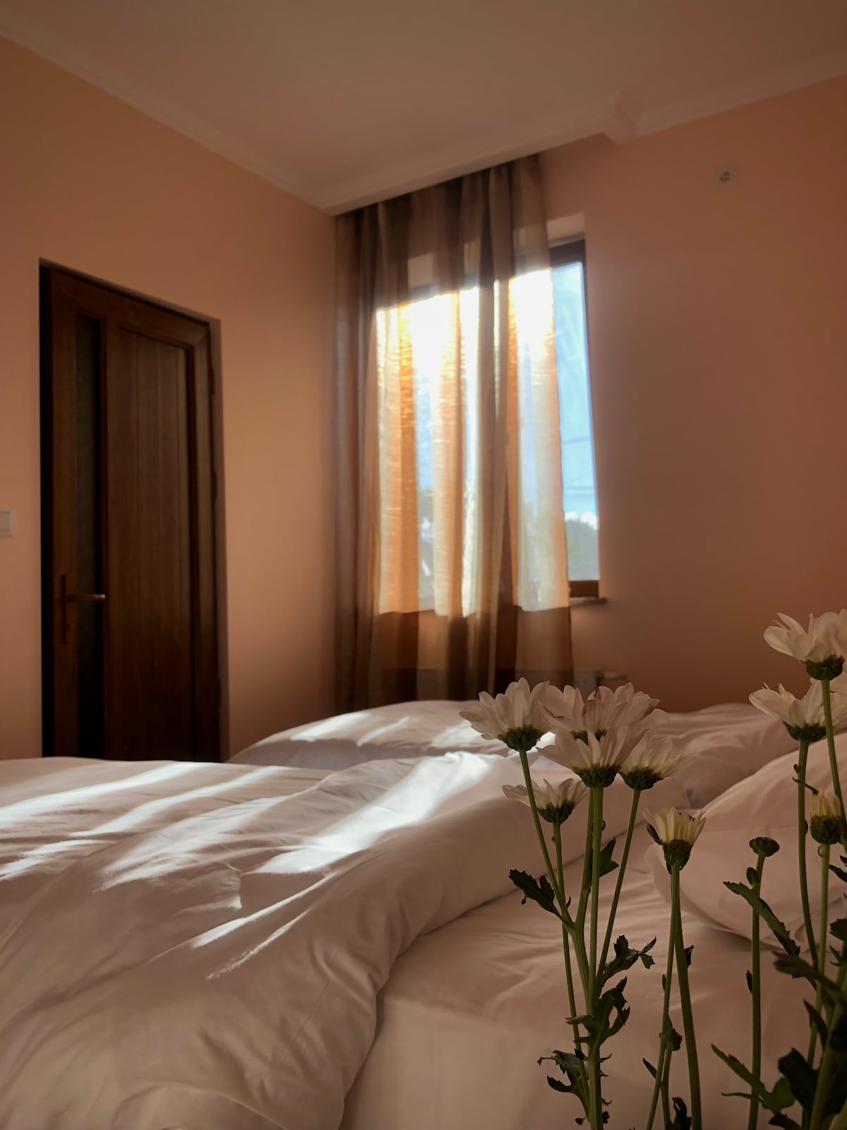 A bright sleeping area is featured with light-colored bedding, neatly arranged on two beds. Natural light filters through a window covered with translucent curtains, illuminating the soft pink walls. A vase with fresh white flowers adds a touch of elegance to the scene.