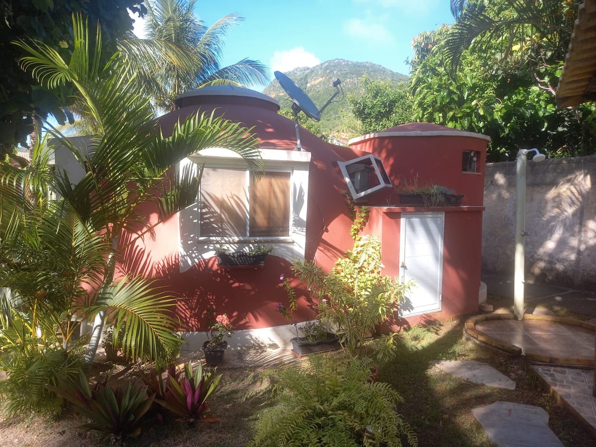 A round, red structure is surrounded by lush greenery, including tall palm trees and colorful plants. A satellite dish is mounted on the roof, while a small garden area features stone pathways leading to the entrance, set against a backdrop of a scenic hillside.