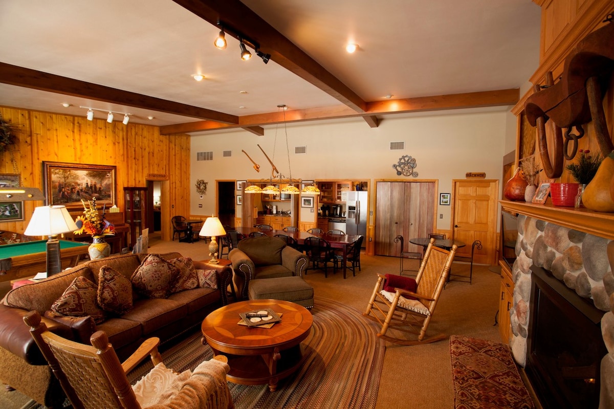 The spacious Great Room features a cozy seating area with a large table and various lounge furniture, accompanied by decorative elements. A stone fireplace serves as a focal point, while views of the fully equipped kitchen and dining area are also visible in the background.
