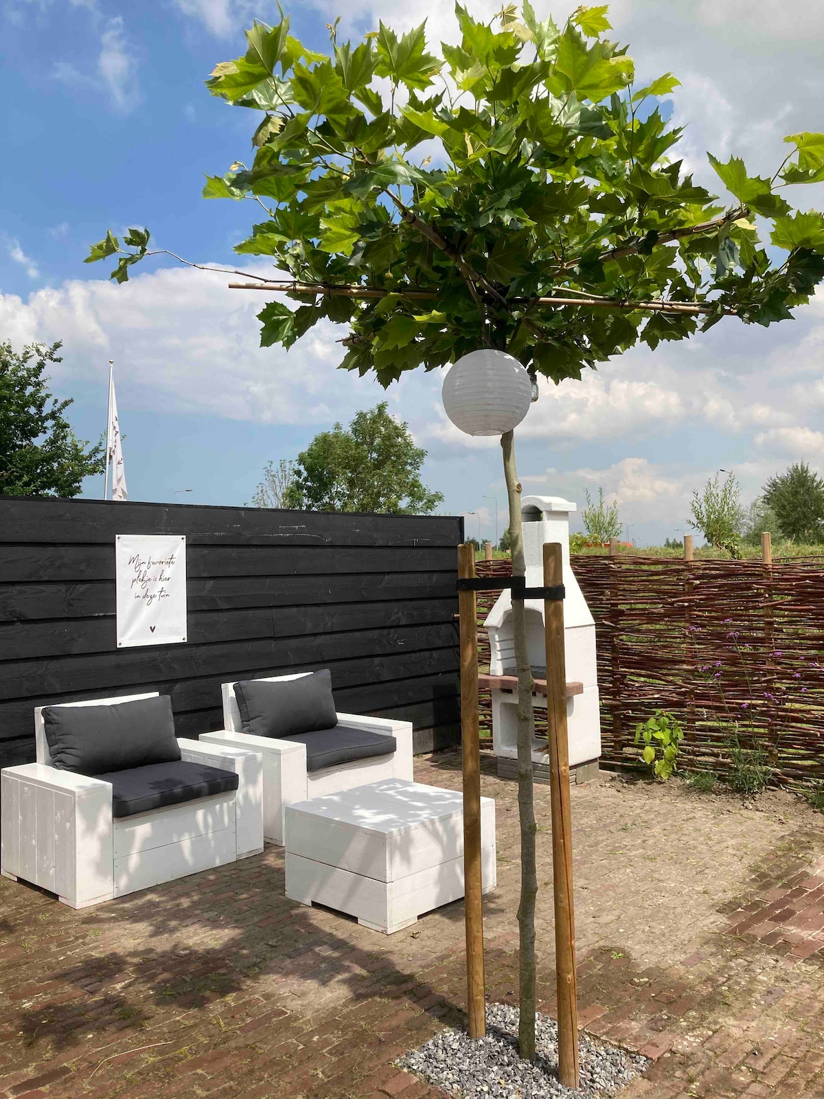 A serene outdoor seating area is depicted, featuring two white chairs with dark cushions. A small tree provides shade, with a decorative lantern hanging from a wooden support. Behind, a simple wooden fence offers privacy, while the ground is paved with outdoor tiles.