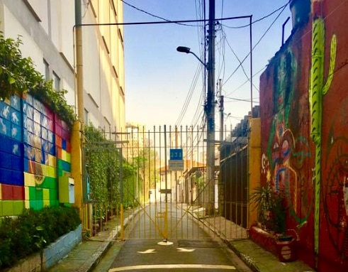 A gated entrance leads into a vibrant street, flanked by colorful murals on both sides. Lush greenery climbs the brick walls, contributing to the lively atmosphere. The clear blue sky is visible above, providing an inviting backdrop to the urban scene.
