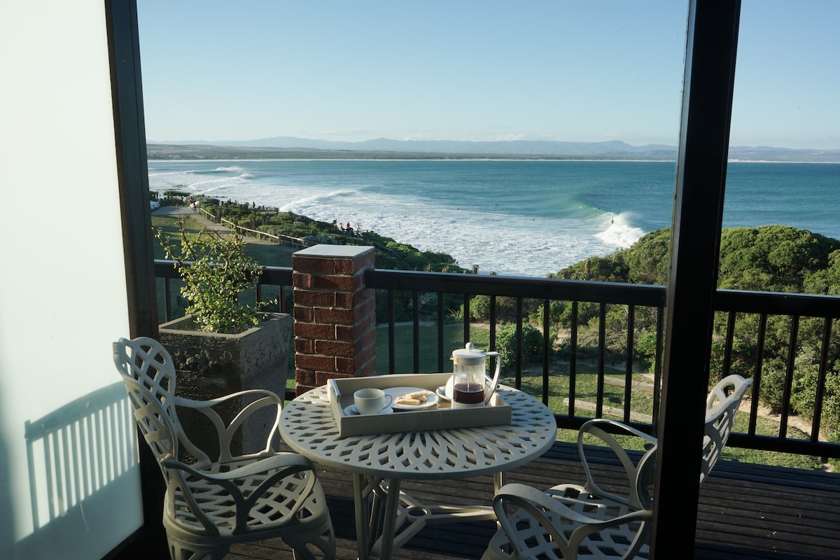A balcony area features a round table with two chairs, set with a coffee pot and a plate of snacks. The stunning ocean view in the background showcases rolling waves and distant mountains under a clear sky.