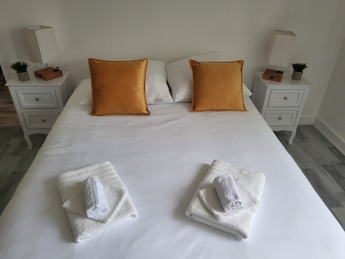 A spacious bed is centered in the room, dressed in crisp white linens. Two decorative pillows in a warm shade of gold are placed at the head of the bed. On either side, white nightstands with small lamps and plants provide a balanced touch.
