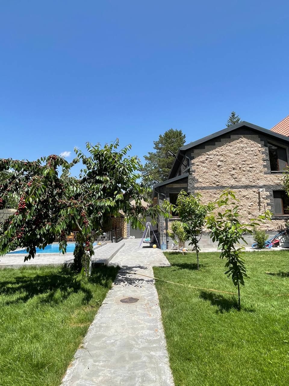 A pathway made of stone leads through a well-kept lawn, bordered by lush trees. The structure, made of natural stone, features large windows and is situated in a sunny outdoor area. A swimming pool is visible in the background, surrounded by greenery.