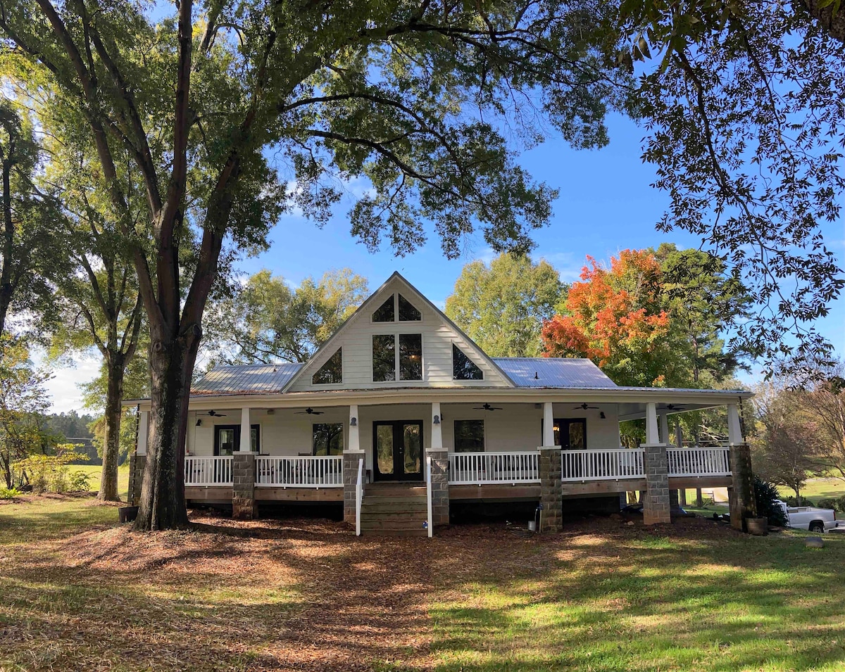 A charming home features a wrap-around covered porch with white railings, surrounded by large trees. The front door is centered and flanked by windows. The roof is pitched, complemented by a clear blue sky and colorful autumn foliage in the background.