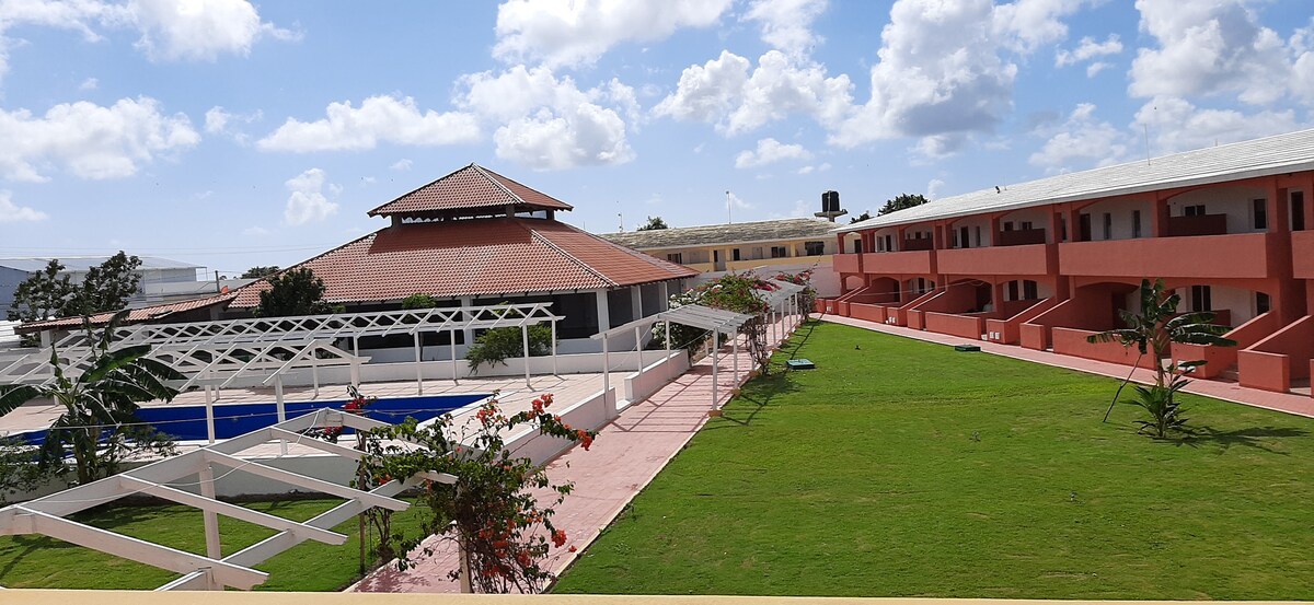 A spacious outdoor area features well-kept greenery and pathways leading to various buildings. A swimming pool is visible in the background, complemented by a shaded pavilion. Surrounding structures exhibit a cohesive architectural style with a combination of vibrant colors and white accents.