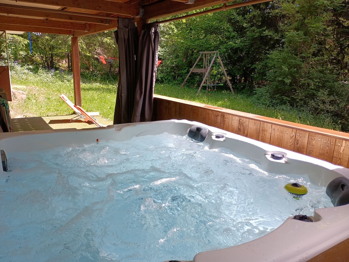 A spacious, private jacuzzi with bubbling water is featured under a wooden shelter. Green foliage surrounds the area, providing a serene setting. Nearby, a wooden swing set and sun loungers are visible, highlighting the outdoor space's tranquility.
