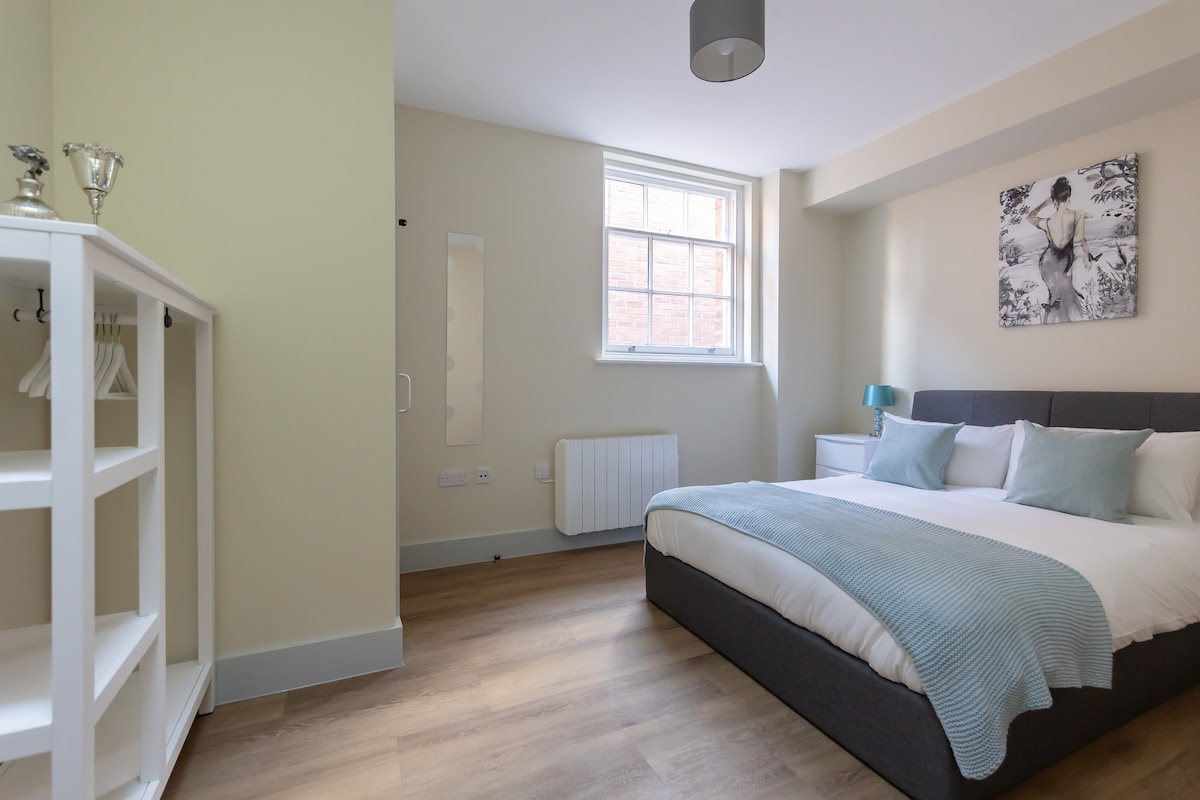 A well-lit bedroom featuring a spacious bed with light bedding and decorative pillows. A wardrobe is positioned against one wall, and a mirror is mounted nearby. Natural light enters through a window, complementing the neutral color scheme and wooden flooring.
