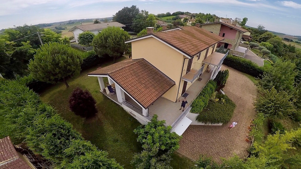 An aerial view of the villa highlights its two-story structure surrounded by lush greenery. A spacious terrace extends from the house, leading to a well-maintained garden. The driveway circles around, providing access to the villa, while panoramic views of the rolling hills can be seen in the background.