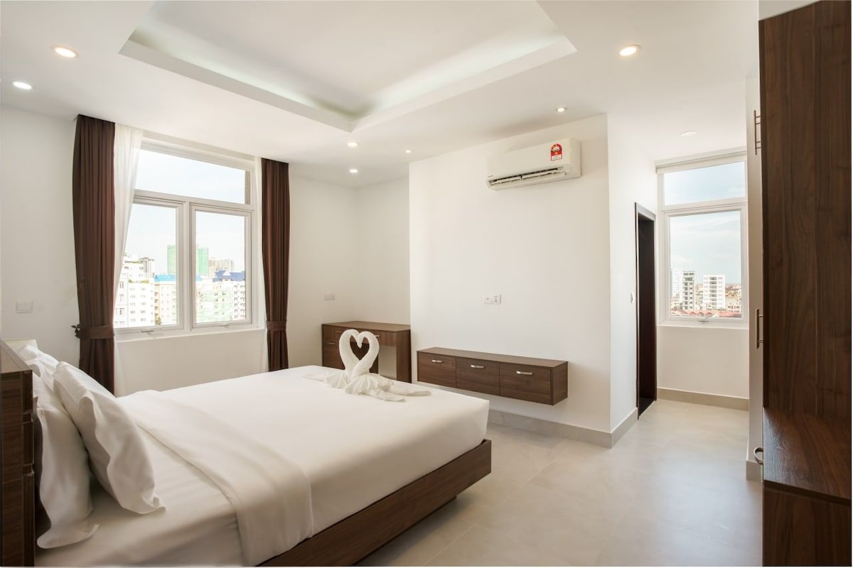 A modern bedroom is presented with light-colored walls and a spacious bed, featuring decorative towel arrangements on the bed. Natural light enters through two windows, and air conditioning is available for comfort. Wall-mounted storage units add functionality to the space.