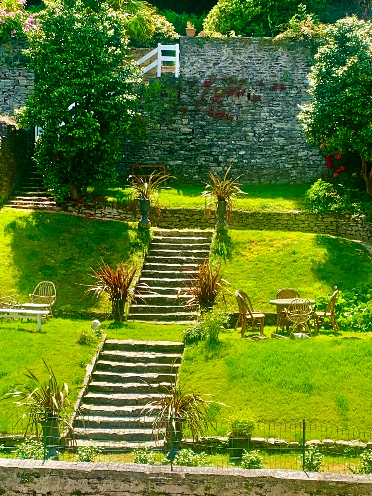 A well-maintained garden is visible, featuring lush green grass and neatly arranged stone steps leading to a higher elevation. Decorative plants are situated along the path, while a small seating area with a table and chairs invites relaxation amidst the natural surroundings.