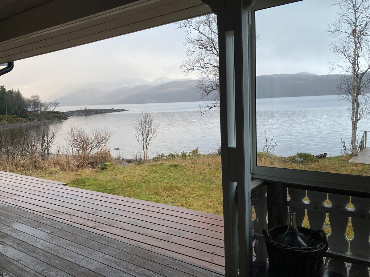 A view from the porch showcases calm waters with reflections of surrounding mountains. The landscape features grassy areas and sparse trees, creating a serene outdoor setting. The wooden deck invites relaxation and enjoyment of the picturesque surroundings.