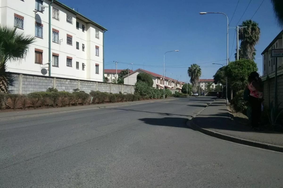 A wide view of a quiet street within a gated community is captured, lined with well-maintained buildings and lush greenery. Palm trees can be seen along the roadside, with clear skies overhead, indicating a calm and sunny atmosphere.