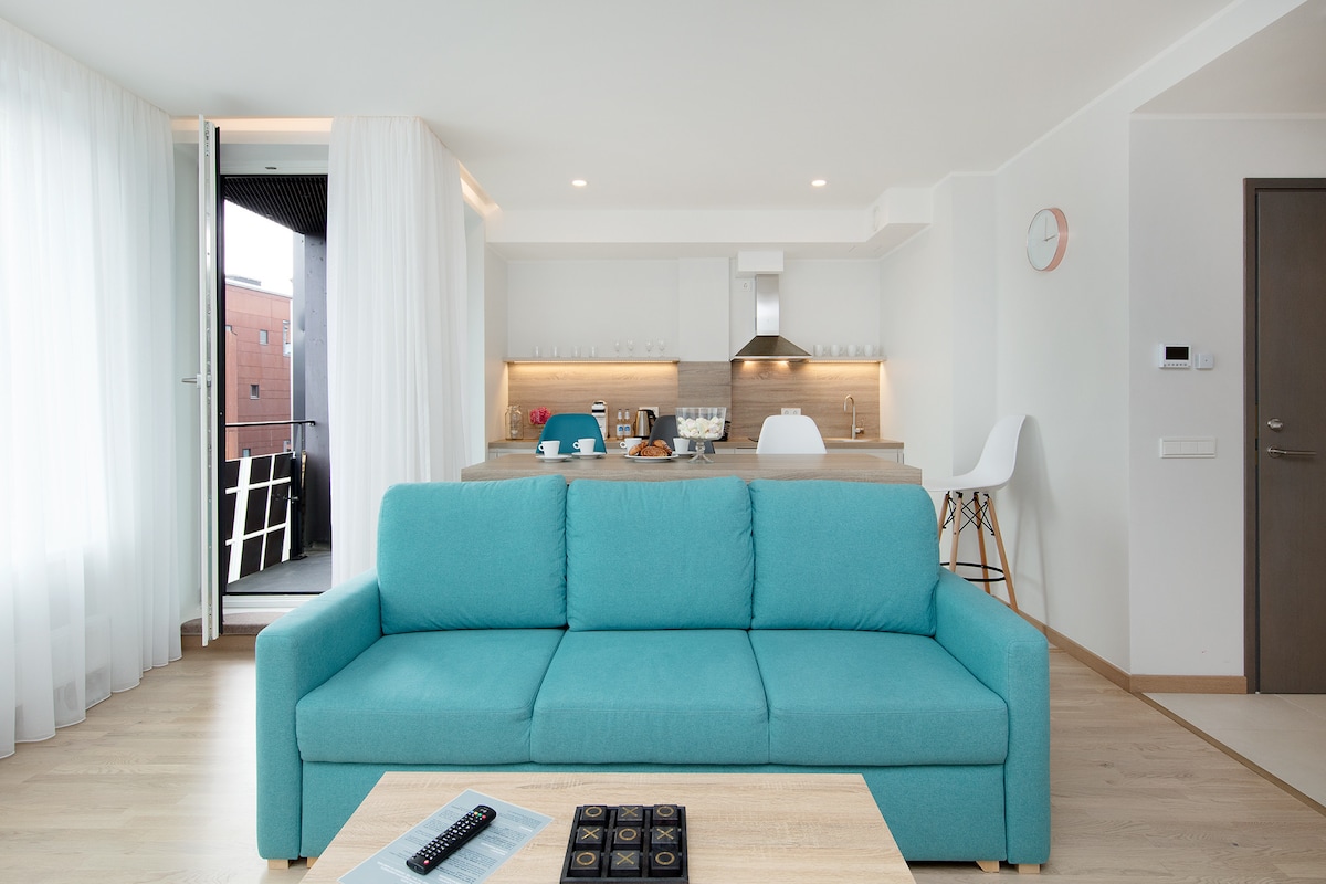 A bright and airy living space is presented, featuring a light blue couch positioned in front of a wooden table. A modern kitchen area with sleek cabinetry is visible in the background, complemented by large windows that open to a balcony.