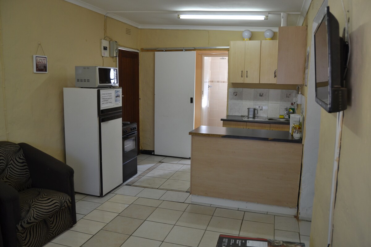 A functional kitchen area is visible, featuring a refrigerator, oven, and countertop. Light-colored cabinetry complements the neutral tile flooring. A black sofa is situated in the corner, while warm overhead lighting illuminates the space. The doorway leads to a bathroom area.