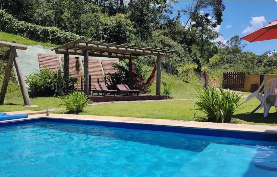 A heated swimming pool is framed by lush greenery, with a pergola offering shaded seating. Two hammocks are suspended near the pergola, creating a relaxed outdoor space amidst the natural landscape.