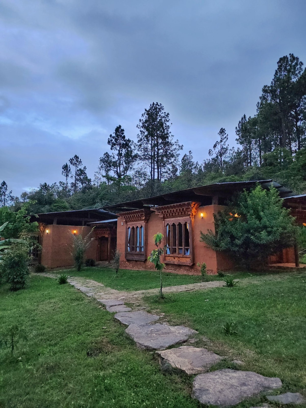 A cozy lodge is surrounded by lush greenery, with stone pathways leading to its entrance. The earthy exterior features traditional architectural elements and warm lighting, while tall trees provide a natural backdrop, contributing to a serene atmosphere in the rural landscape.