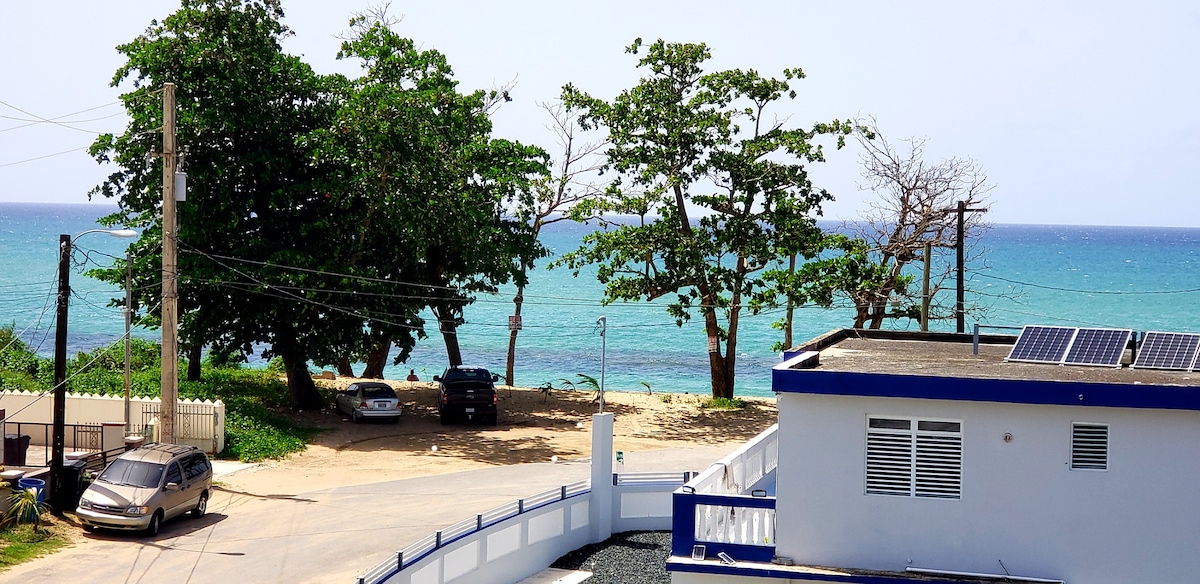 A scenic ocean view is displayed, featuring a sandy beach framed by lush green trees. The gentle waves of the sea create a calming backdrop, while a parking area can be seen nearby. Solar panels are visible on the roof of the adjacent building.