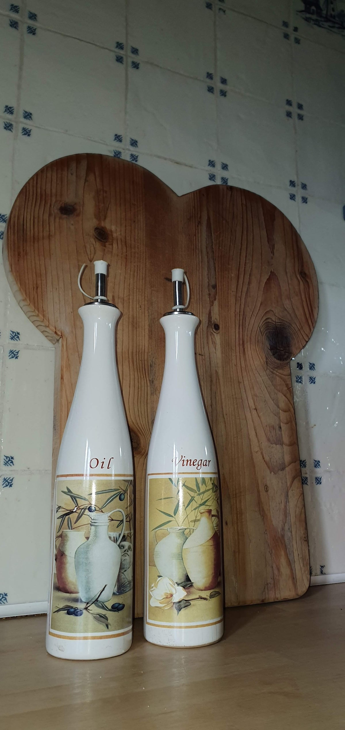 Two elegant glass bottles labeled 'Oil' and 'Vinaigra' are displayed next to a heart-shaped wooden cutting board. The background features white tiles with a blue pattern, creating a warm kitchen ambiance.