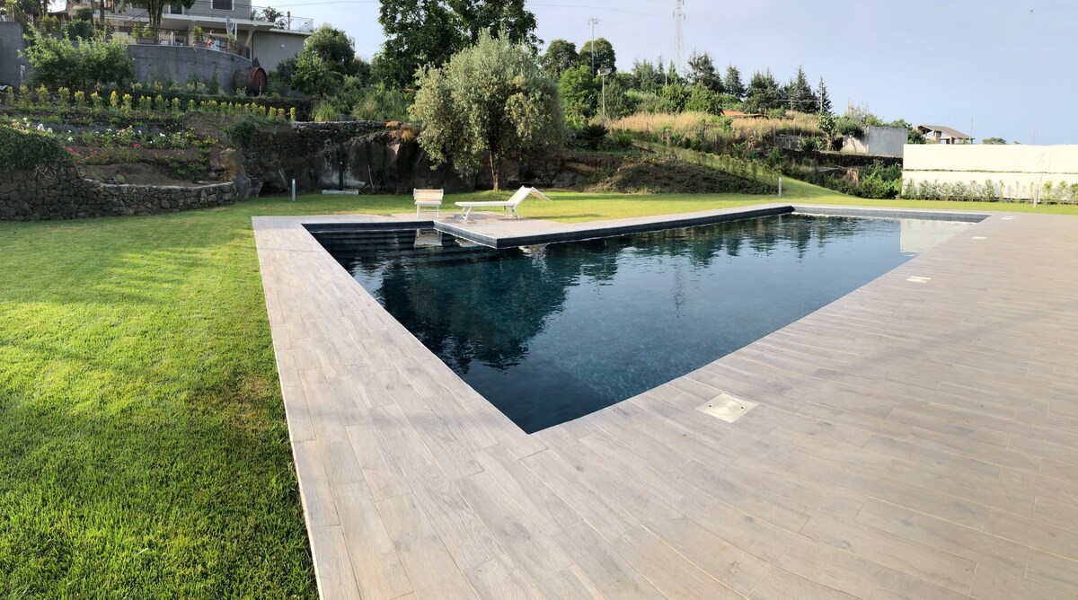 A spacious outdoor salt-water pool is situated within a well-maintained garden, bordered by lush greenery. Surrounding the pool, a wooden deck provides ample space for relaxation. The view displays terraced landscaping with vibrant flower beds in the background, enhancing the serene outdoor setting.