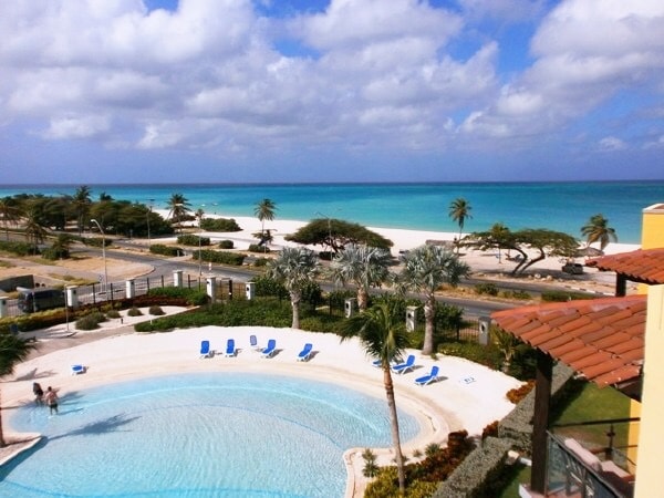 A panoramic view showcases a serene beach and azure waters, framed by lush palm trees. Below, a curving pool reflects the sky, while lounge chairs are positioned around the pool area. The ocean extends in the distance, offering a peaceful coastal atmosphere.