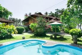 A charming resort view is presented, featuring a refreshing pool surrounded by lush greenery. The wooden structure is partially visible in the background, nestled among flowering plants and trees. A shaded area with a sun umbrella adds to the tranquil outdoor space.