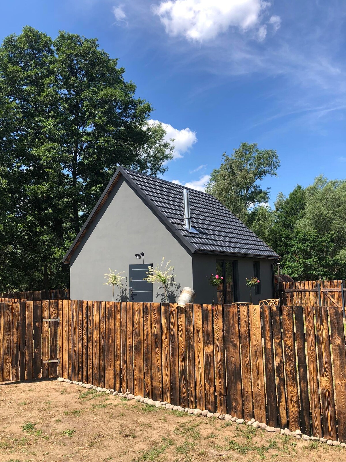 A charming cabin is framed by a rustic wooden fence, surrounded by greenery. The structure features a sloped roof and is enhanced by flower boxes near the windows. A clear blue sky with scattered clouds complements the peaceful rural atmosphere.