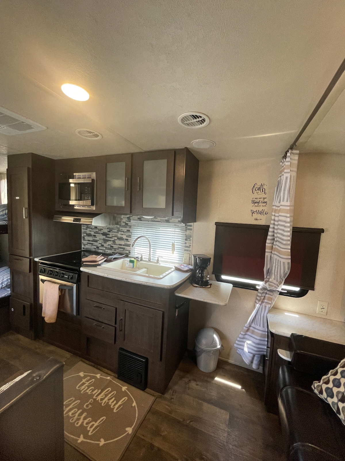 The compact kitchen area features dark cabinetry, a microwave, and a stove. A small sink is positioned beneath a window with shades. A coffee maker and trash bin are present, while light wood flooring adds warmth to the space.