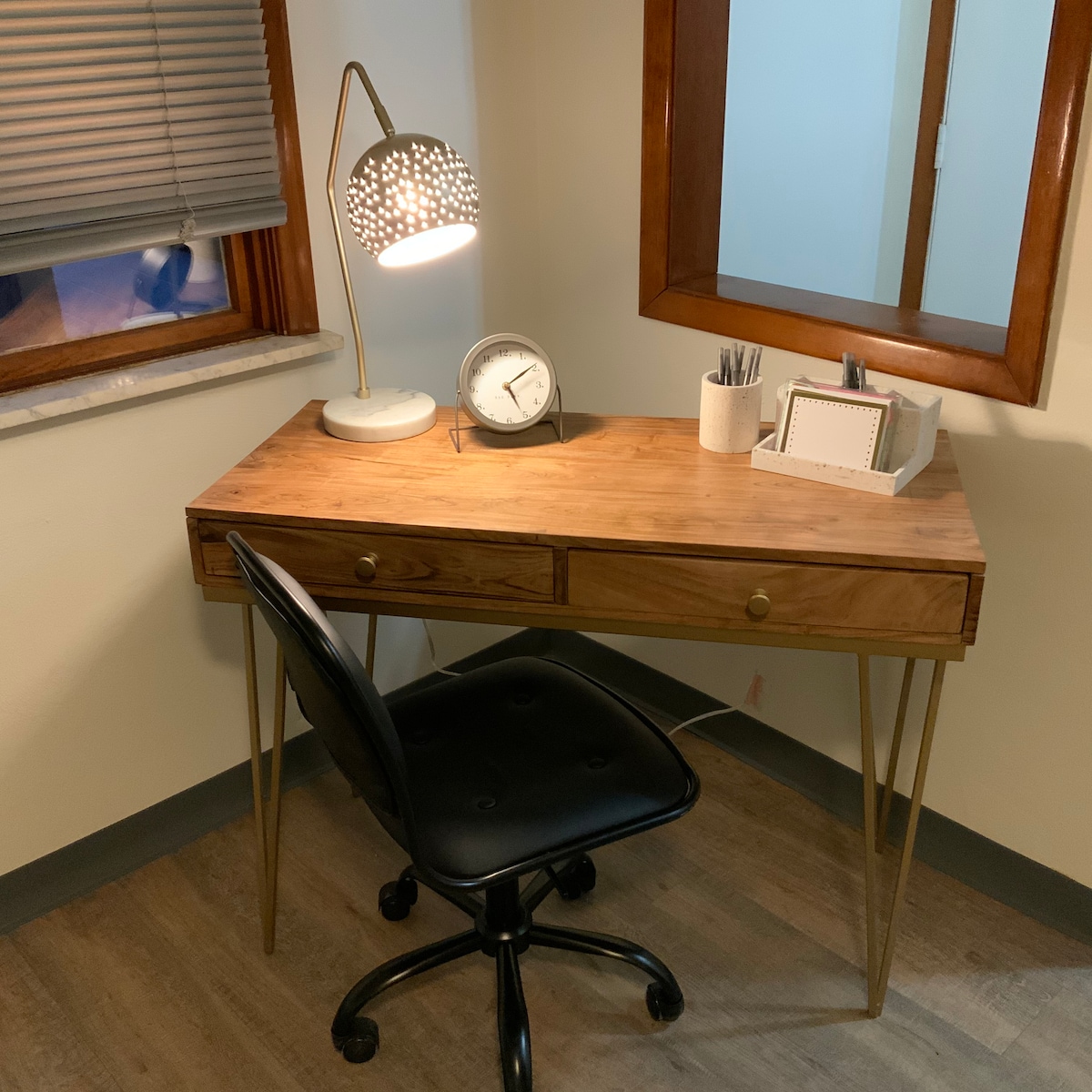 A wooden desk with two drawers is positioned against a wall. A modern desk lamp provides light, accompanied by a clock and writing utensils organized in a holder. A black rolling chair is situated in front of the desk, and the room features a window with blinds.