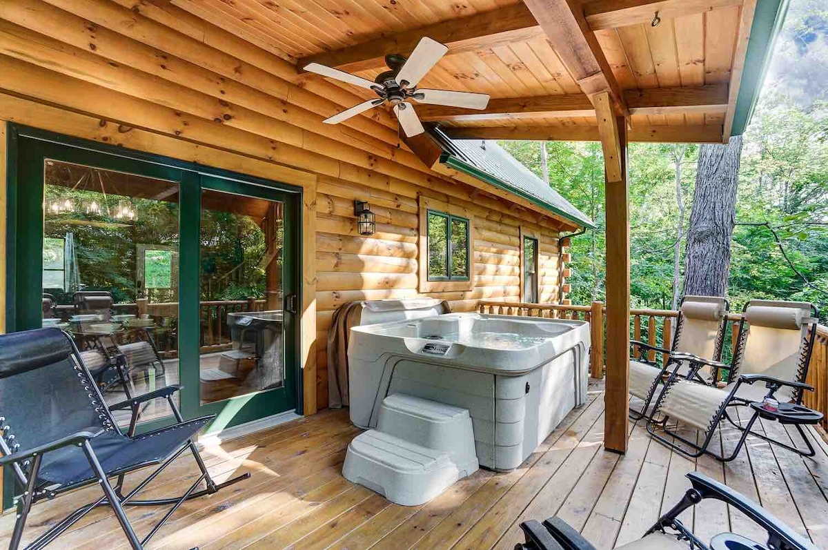The outdoor deck features a hot tub designed for six, surrounded by two Zero Gravity chairs and a ceiling fan for comfort. Sliding glass doors offer a view into the cabin, framed by wooden siding and lush greenery.