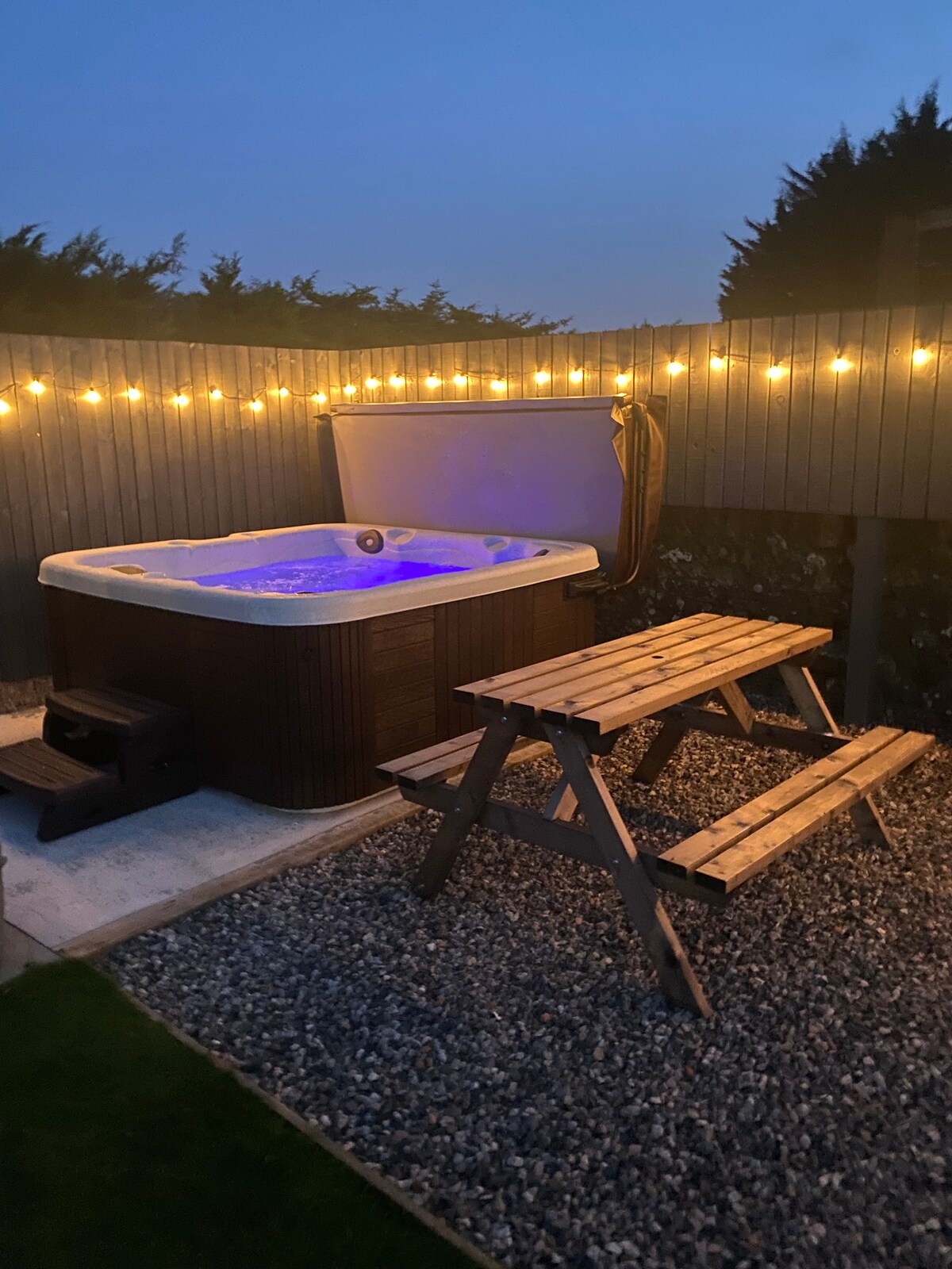A well-lit outdoor area features a hot tub with calming purple illumination. Nearby, a wooden picnic table is surrounded by a gravel surface and bordered by a fence adorned with string lights, creating a cozy and inviting setting for relaxation.
