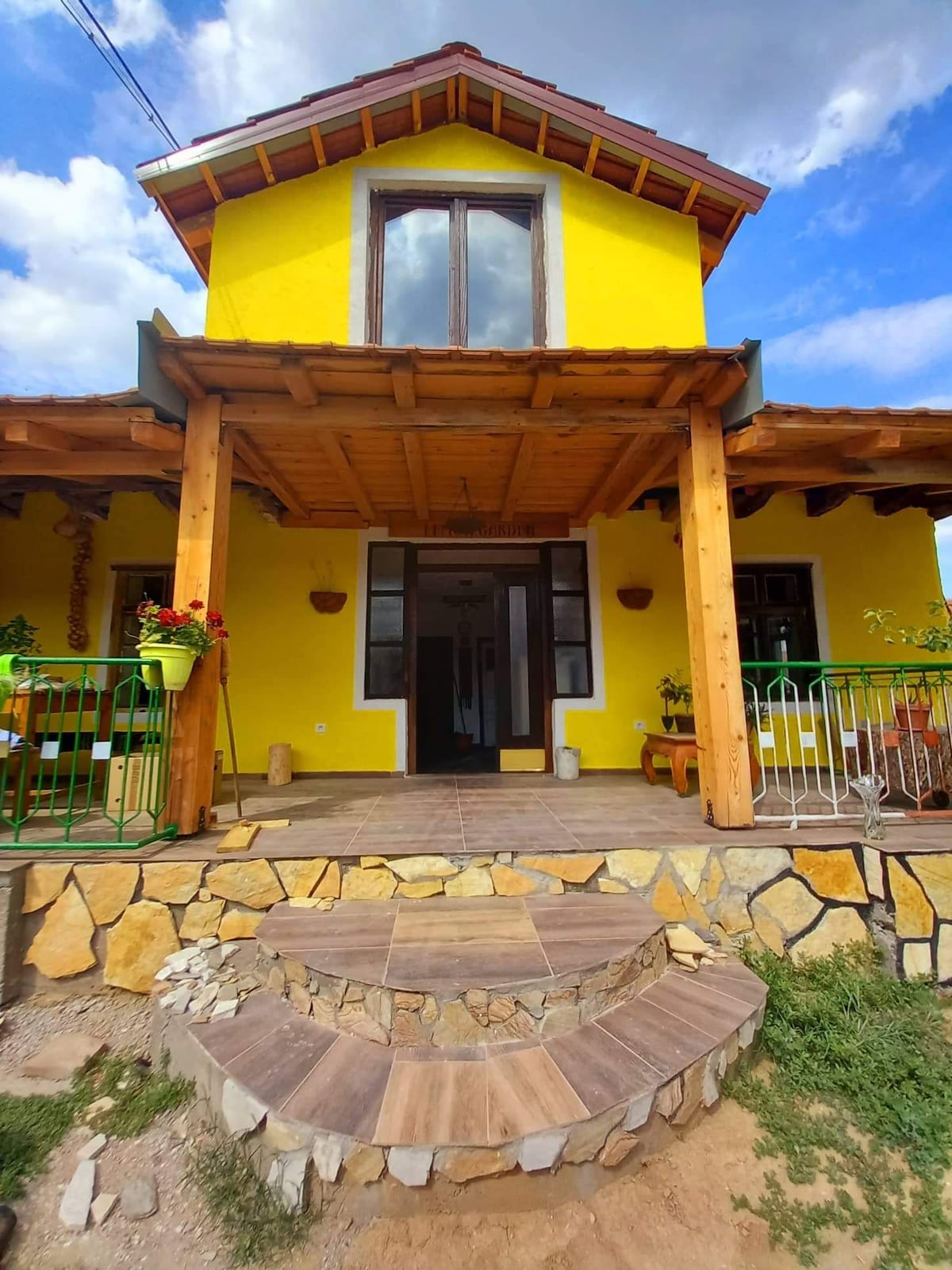 The exterior of a vibrant yellow house is presented, featuring a wooden porch supported by sturdy beams. Stone steps lead to the entrance, while potted plants adorn the railing, adding a touch of greenery. Large windows allow natural light to fill the inviting space.