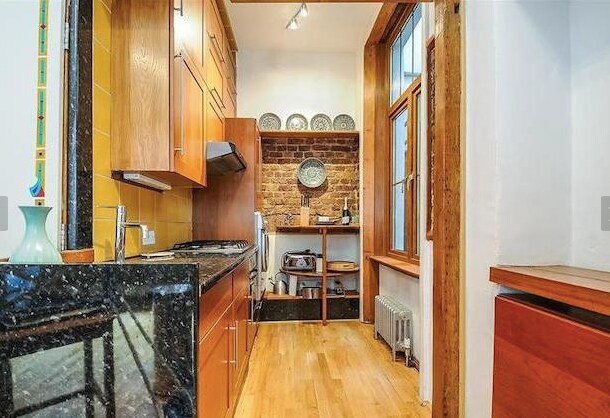 A well-equipped kitchen features wooden cabinetry and a granite countertop. Exposed brick adds character, while a window allows natural light to illuminate the space. Shelves hold dishware, and modern appliances, including a gas hob, are integrated into the design.