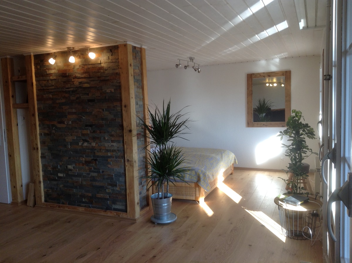 A serene room features natural wooden flooring and a textured stone wall accent. A spacious bed is positioned against the wall, with sunlight filtering in through a window. Decorative plants enhance the atmosphere, complemented by soft overhead lighting.