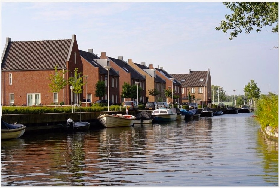 A serene waterfront scene features a row of modern houses set along a calm canal. Various boats are moored beside the grassy bank, while small trees provide greenery. The clear sky reflects off the water, enhancing the peaceful ambiance of the neighborhood.