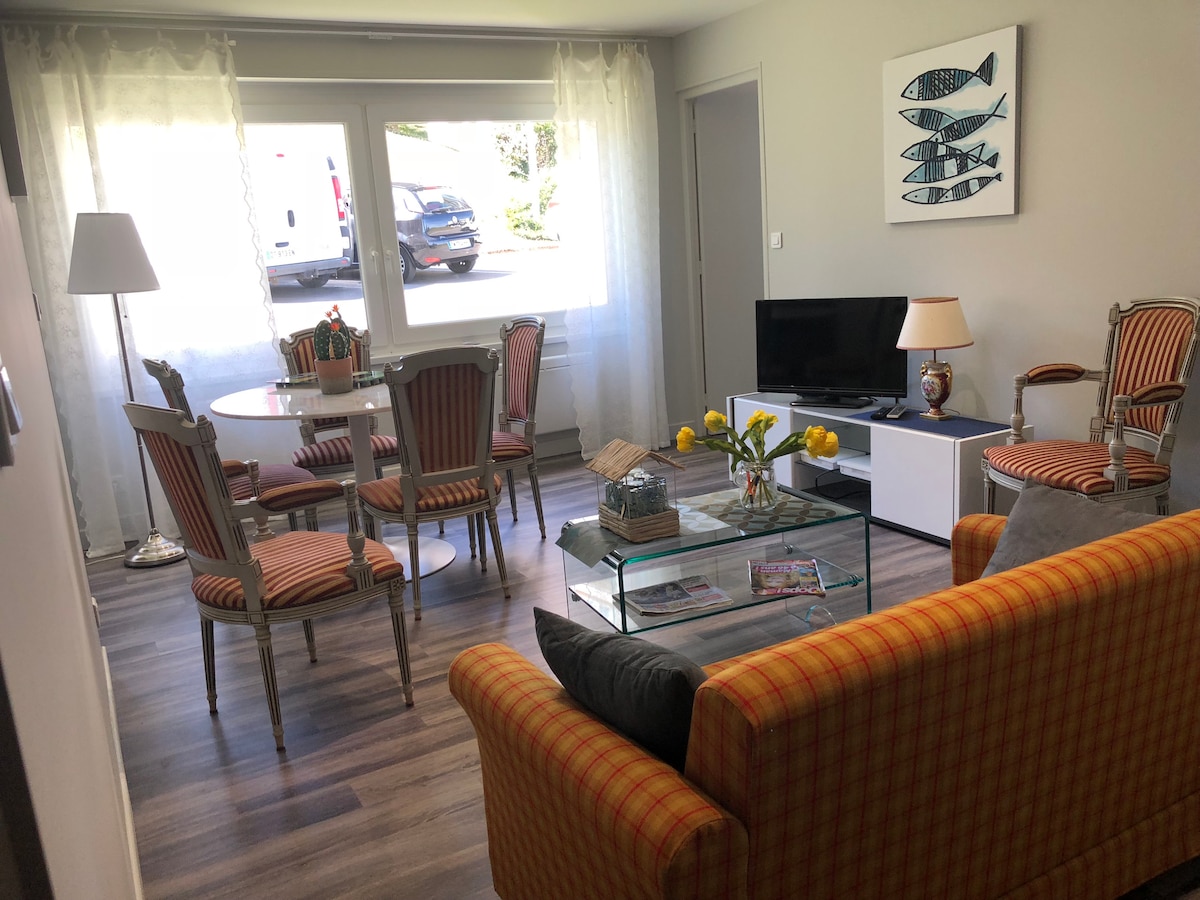 A light-filled living area features a comfortable sofa with a striped pattern and a glass coffee table. A dining set with six striped chairs is visible, along with a lamp in the corner. A piece of wall art depicting fish adds a decorative touch to the space.