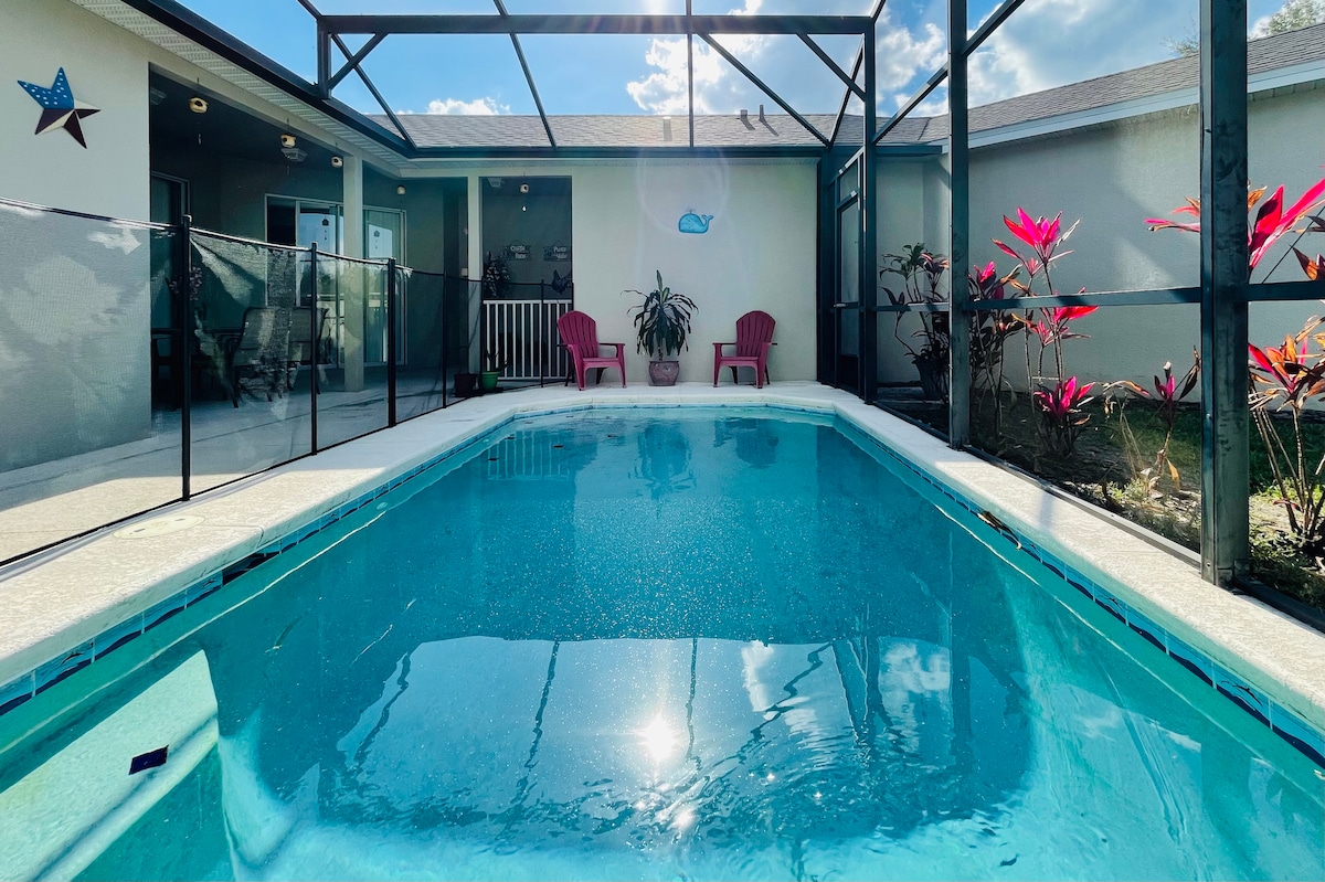 A private screened pool is showcased with clear blue water, surrounded by a light-colored patio. Colorful plants are positioned around the area, and two red chairs are placed near the poolside. The background features a residential home with an open area and additional seating visible.