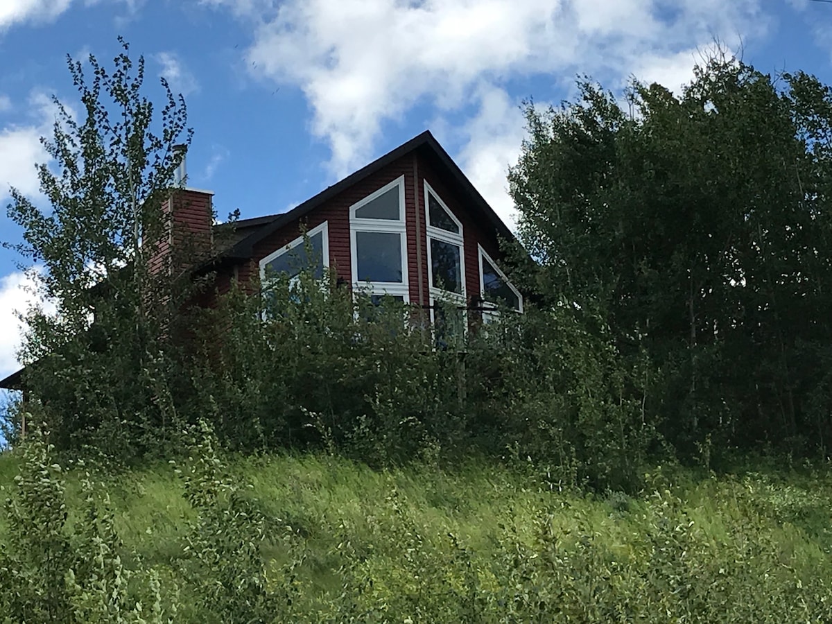 The red cabin is nestled among greenery, showcasing large windows that offer glimpses of the outdoors. The sloped roof complements the natural landscape, with clouds visible in the sky above, creating a serene backdrop.