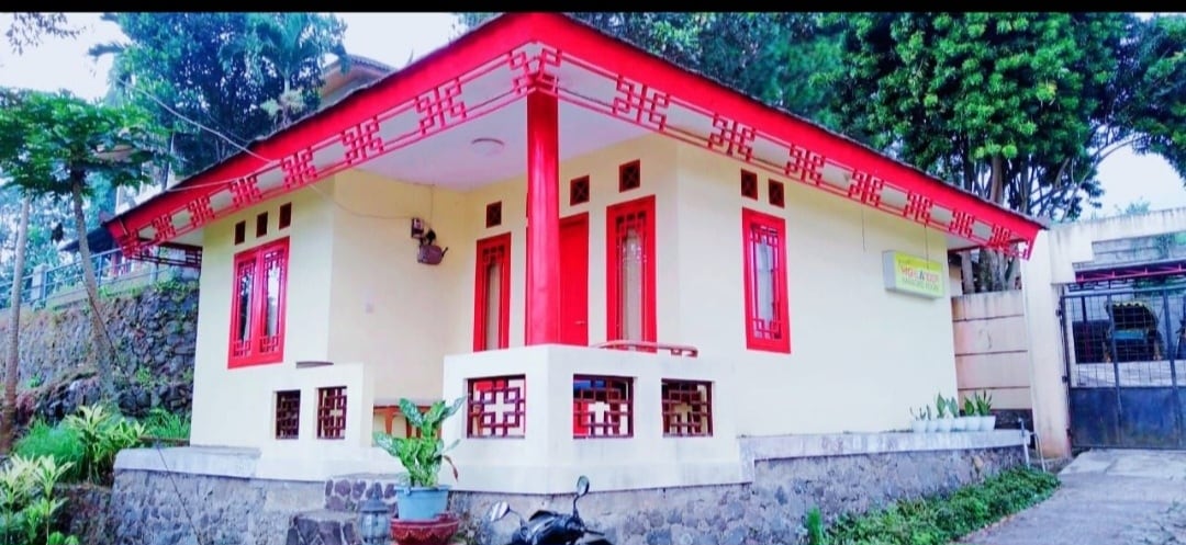 A quaint house is presented with a vibrant red roof and complementary red detailing around the windows. The structure is surrounded by greenery, with decorative plants placed near the entrance. A stone pathway leads to the home, enhancing the welcoming atmosphere.