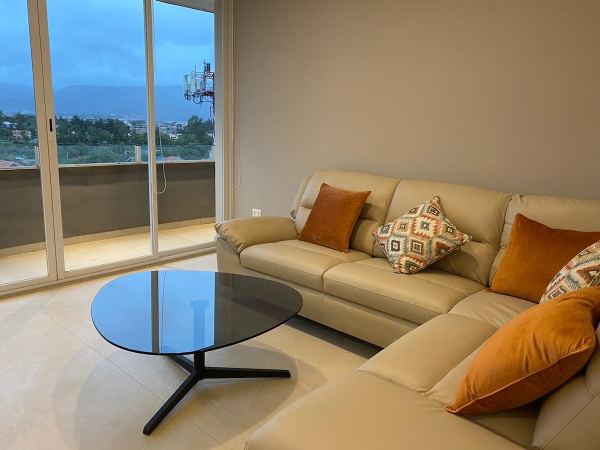 A modern living area is displayed, featuring a light-colored leather sofa adorned with decorative cushions. A sleek black coffee table is positioned at the center. Large glass doors open to a view of the outdoors, allowing natural light to fill the space.