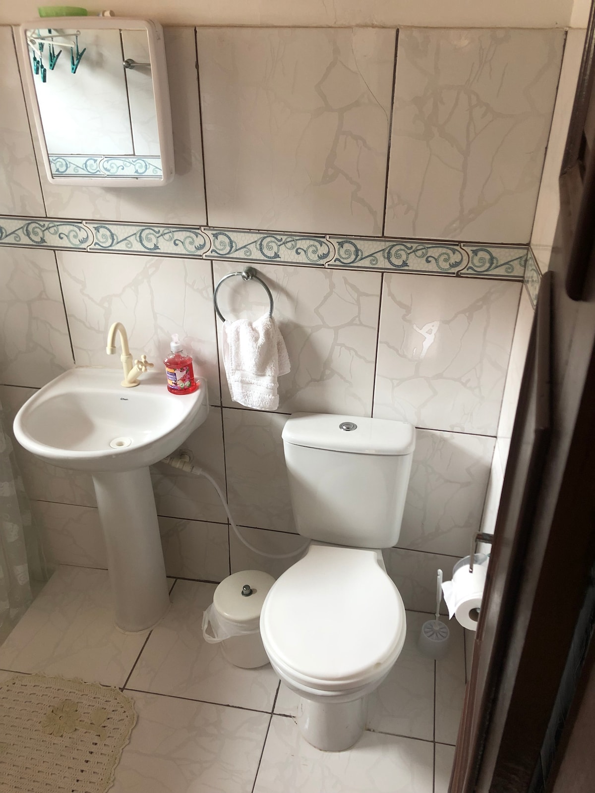 The bathroom features a ceramic sink with a mounted mirror above. A modern toilet is positioned adjacent to the sink. The walls are tiled with a subtle marble pattern, and a towel hangs from a nearby rack. Natural light filters in through a window.